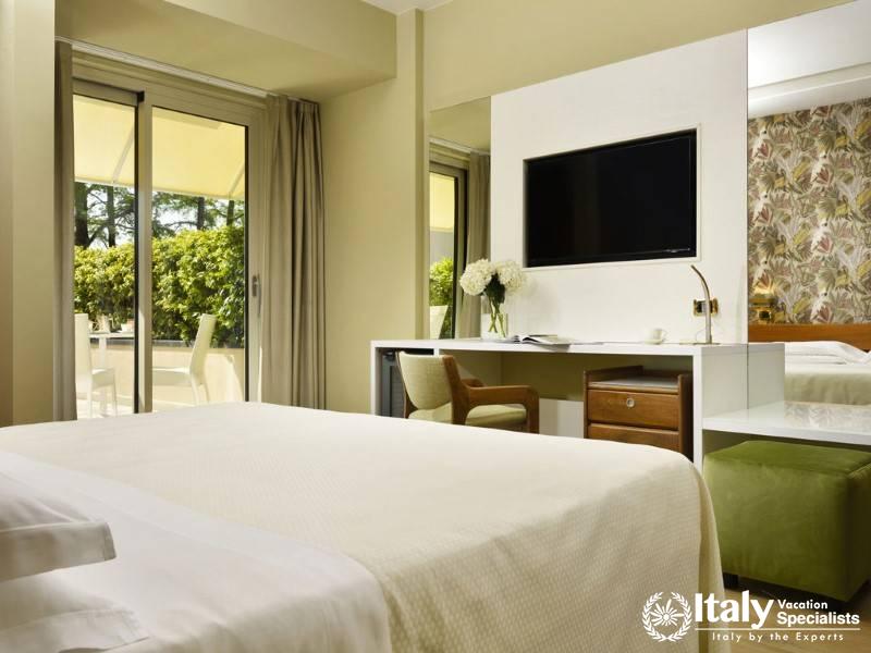 A well-lit, stylish superior room with a cozy bed and private balcony, overlooking lush greenery.