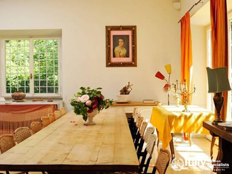 Sunlit dining area with a long wooden table and a portrait on the wall.