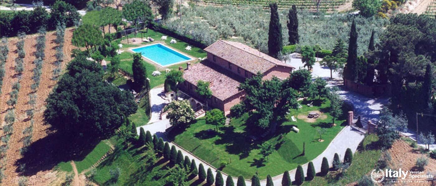 An aerial view of a grand Tuscan area surrounded by gardens, a pool, and vineyards.