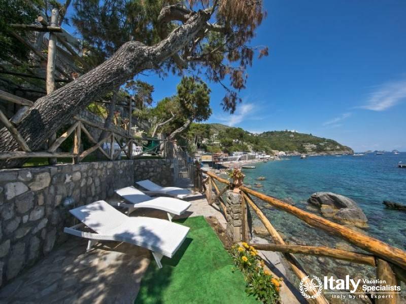 Additional view of loungers by the clear blue sea and rocky coastline.